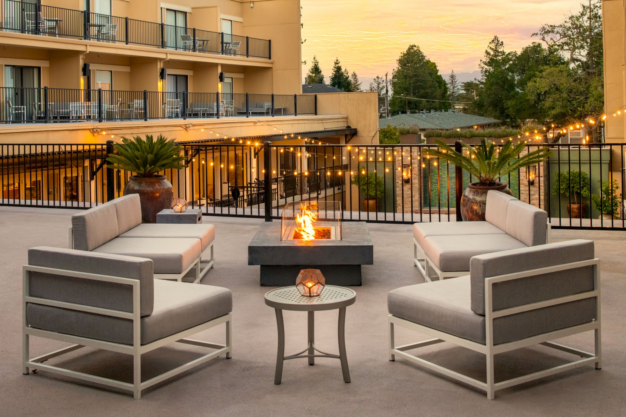 The image features an outdoor lounge area with modern seating, a fire pit in the center, fairy lights, and a background of a building and trees at sunset.