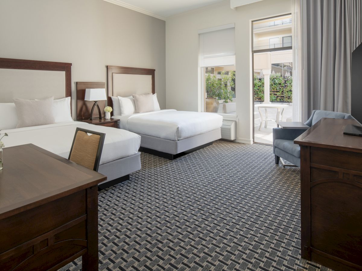 The image shows a tidy hotel room with two double beds, a desk with a chair, a TV on a dresser, and a large window with curtains.