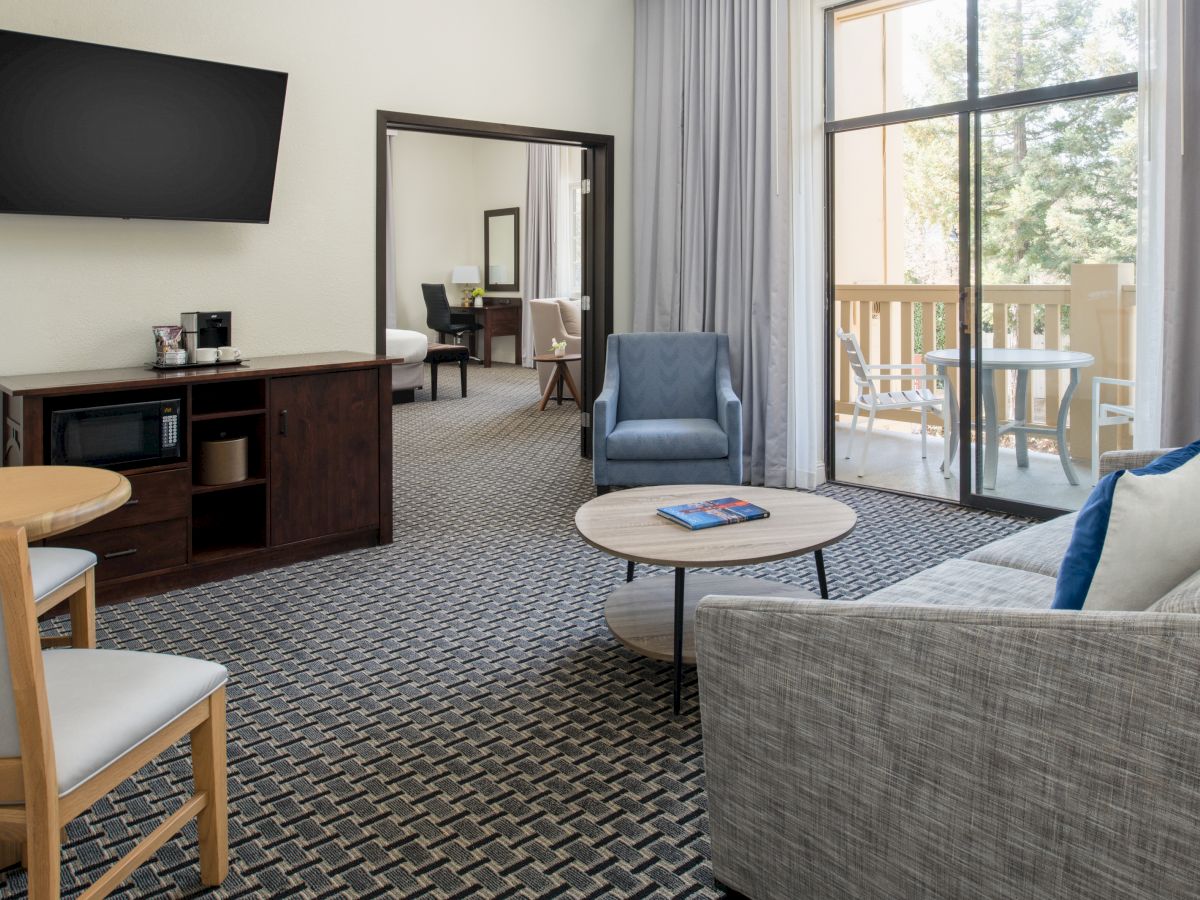 A modern living room with a sofa, armchair, coffee table, wall-mounted TV, and large window. There is a dining area and a door leading to another room.