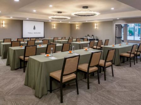 A conference room with rows of tables and chairs, a screen displaying "Toll House", and refreshments on the tables. The room is well-lit.