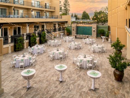 An outdoor event space with round tables and white chairs, tall cocktail tables, string lights above, a water feature, and a sunset background.