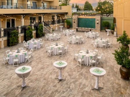 The image shows an outdoor event space with round tables and chairs, high tables, string lights, greenery, and a water feature on the far wall.