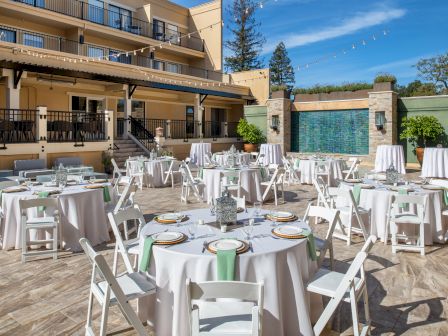 An outdoor dining area is set up with round tables, white tablecloths, green napkins, and white chairs. A multi-story building and fountain are visible.