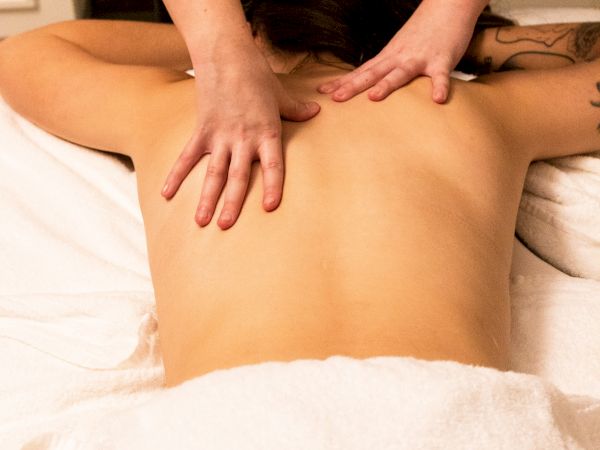 The image shows a person receiving a back massage, with a masseuse applying pressure to the upper back while the person lies on a white towel-covered table.