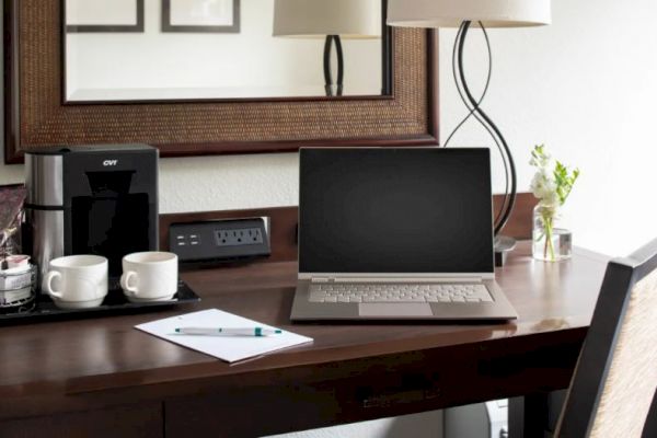 A tidy workspace with a laptop, two cups, a coffee maker, a power outlet with USB ports, a pad and pen, a vase with flowers, and a lamp under a mirror.