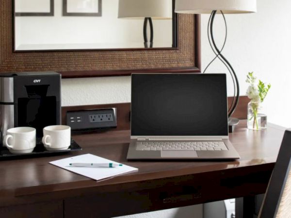 A tidy workspace with a laptop, two cups, a coffee maker, a power outlet with USB ports, a pad and pen, a vase with flowers, and a lamp under a mirror.