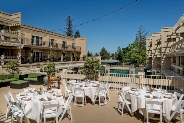 Outdoor dining area with round tables and white chairs, set up with tableware. Surrounded by buildings and greenery under a clear blue sky.