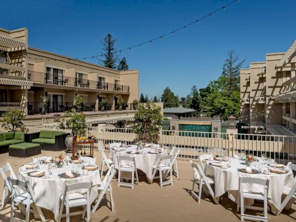 Outdoor dining area with round tables and white chairs, set up with tableware. Surrounded by buildings and greenery under a clear blue sky.