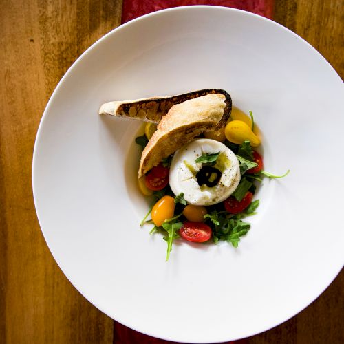 A beautifully plated dish featuring sliced bread, a poached egg on greens, cherry tomatoes, and garnish on a white plate.