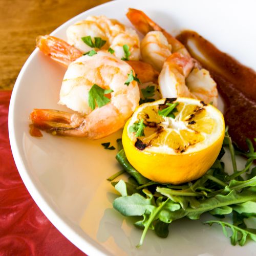 The image shows a white plate with shrimp, a grilled lemon half, a bed of greens, and a red sauce on the side, placed on a wooden table.