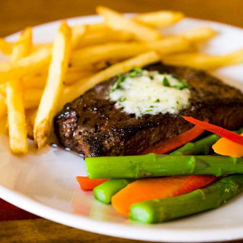 A plate with steak topped with herb butter, french fries, and steamed vegetables including asparagus, carrots, and red bell pepper.