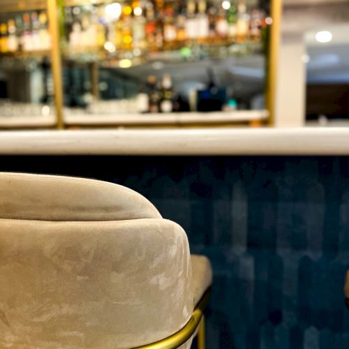 Comfortable upholstered bar stool with a golden frame in front of a stocked bar with various bottles and glassware on shelves in the background.