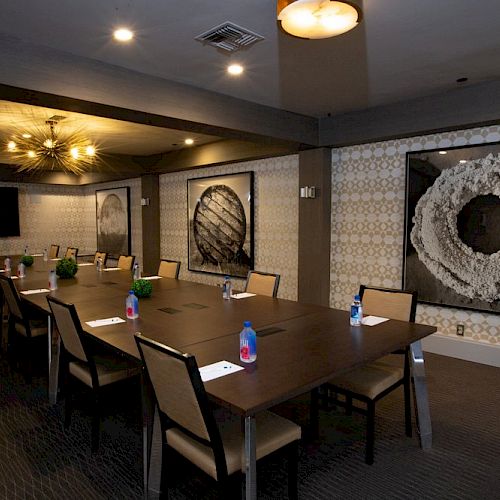 This image shows a modern conference room with a long table, chairs, notebooks, and water bottles, featuring wall art and a flat-screen TV.