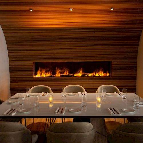 A modern, elegant dining table set for eight, located in front of a lit, narrow fireplace with a wooden backdrop, creating a cozy ambiance.