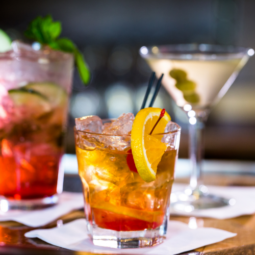 Three cocktails with different garnishes are displayed on a bar; one with citrus slices, one with mint and cucumber, and one with olives.