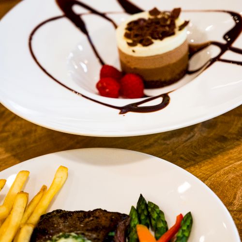 The image shows two plates: one with steak, fries, asparagus, and vegetables; the other with a dessert, garnished with raspberries and chocolate sauce.