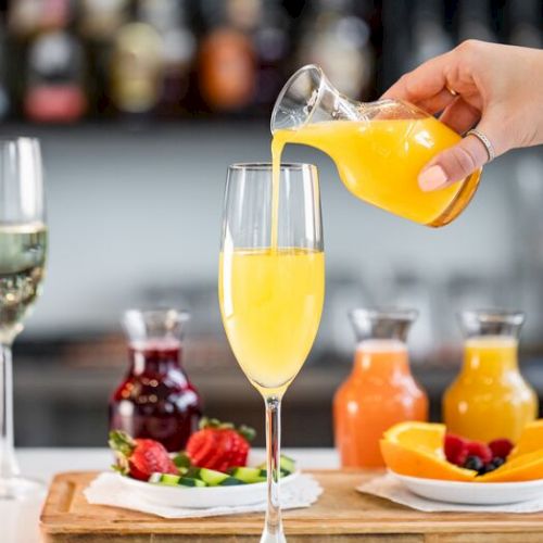 A bartender pours orange juice into a champagne glass on a table with fruit and bottles of mixers.