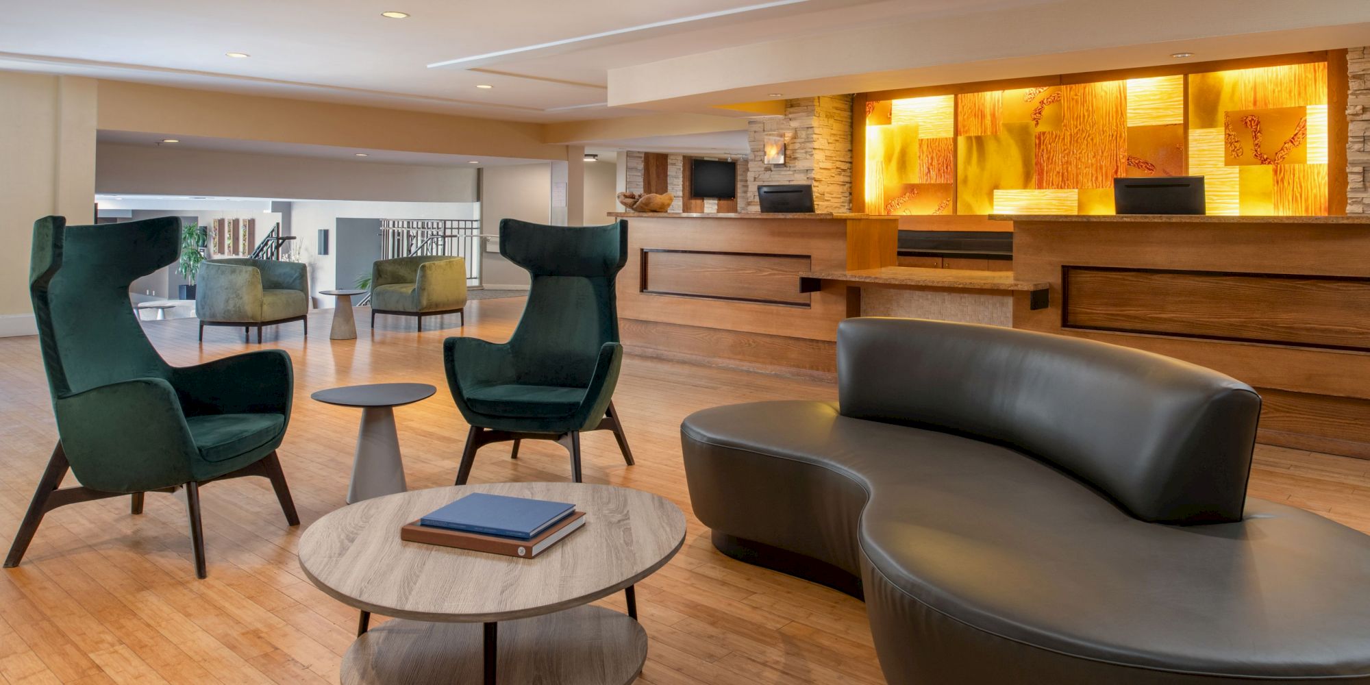 A modern lobby with stylish chairs, a curved sofa, wooden flooring, a reception desk with warm lighting, and a small table with a book on it.