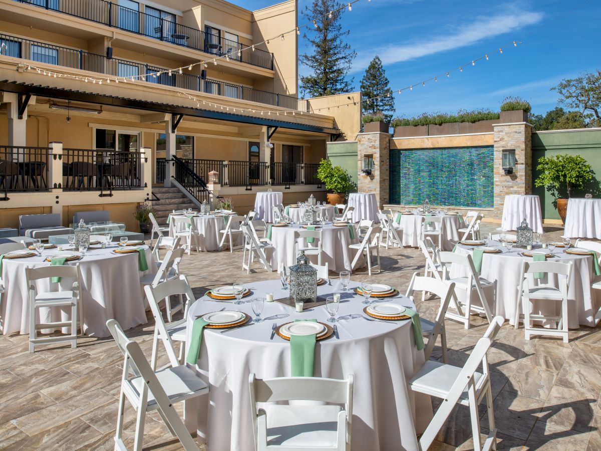 An outdoor patio setup for an event with round tables, white tablecloths, and chairs arranged neatly. A fountain and string lights are visible.