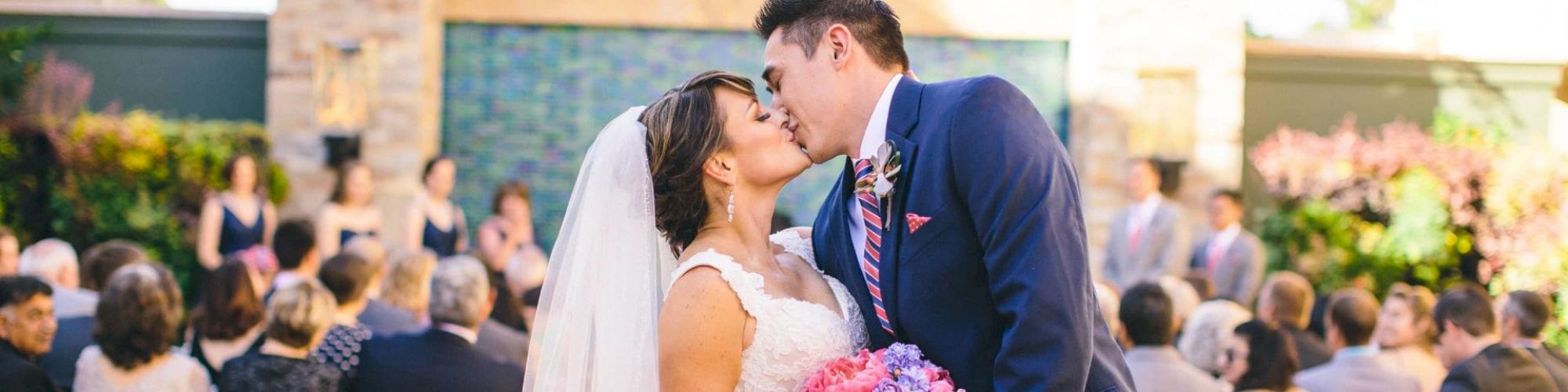 The image shows a bride and groom sharing a kiss at their wedding ceremony, with guests seated and the bridal party in the background.