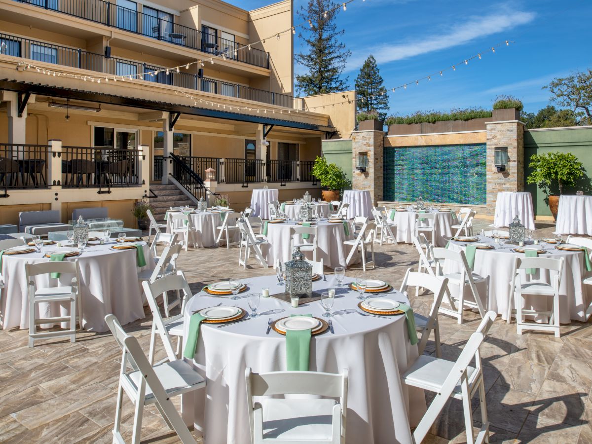 An outdoor patio setup for an event, featuring round tables with white tablecloths, white chairs, and place settings, in front of a water feature.