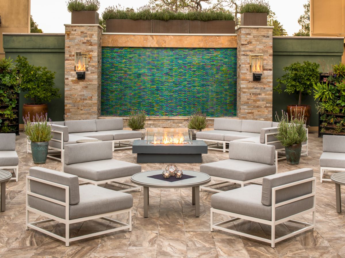 This image shows an outdoor seating area with modern furniture, a fire pit in the center, and a water feature on the back wall with plants around.