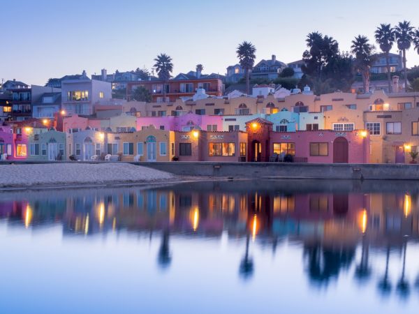 A serene waterfront scene with colorful buildings reflecting on calm water, with palm trees in the background and lights creating a tranquil ambiance.
