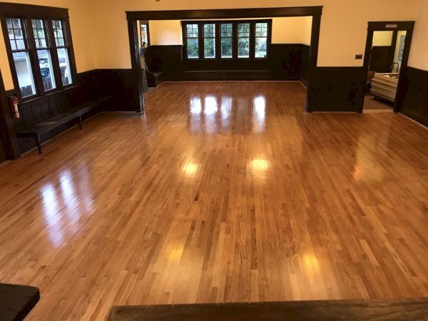 The image shows an empty room with wooden flooring, dark wooden trim, multiple windows, and some benches along the walls.
