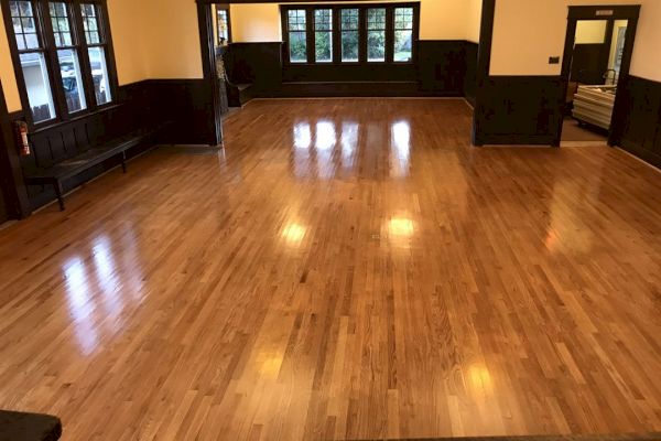 The image shows an empty room with wooden flooring, dark wooden trim, multiple windows, and some benches along the walls.