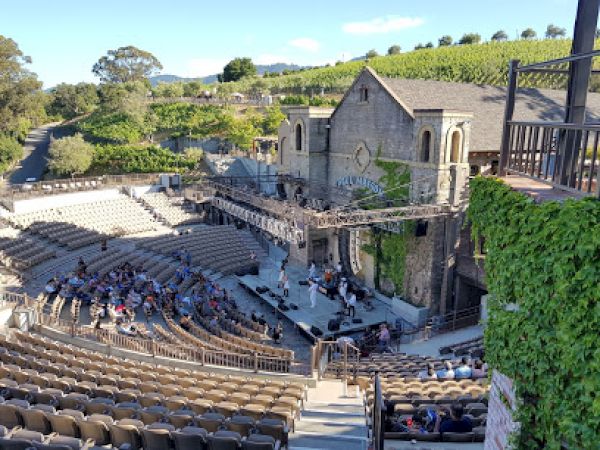 The image shows an outdoor amphitheater with tiered seating and a stage set in front of a stone building with greenery in the background.