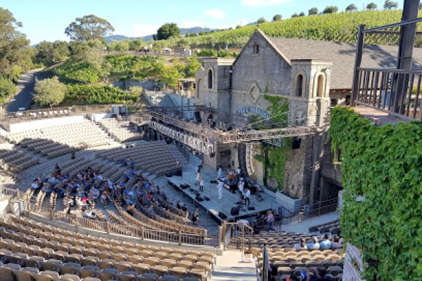 The image shows an outdoor amphitheater with tiered seating and a stage set in front of a stone building with greenery in the background.