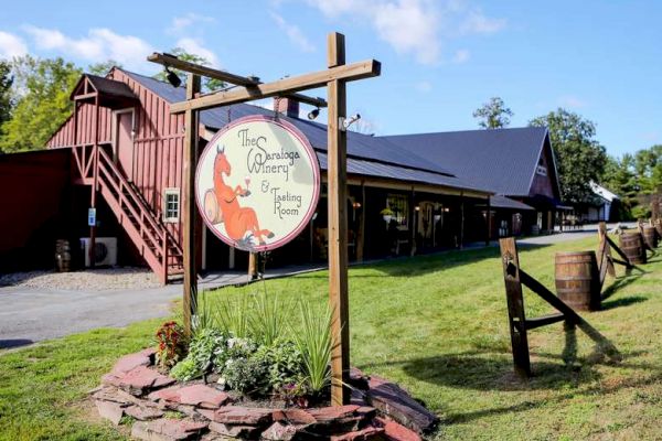 The image shows a rustic winery with a sign that reads "The Saratoga Winery & Tasting Room" in front of a barn-style building and green, landscaped grounds.