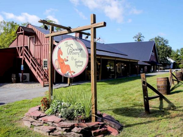 The image shows a rustic winery with a sign that reads "The Saratoga Winery & Tasting Room" in front of a barn-style building and green, landscaped grounds.