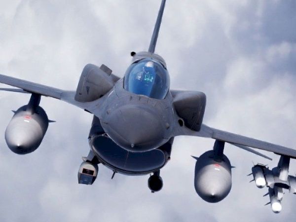 The image shows a front view of a military fighter jet flying with clouds in the background, equipped with various weapons.