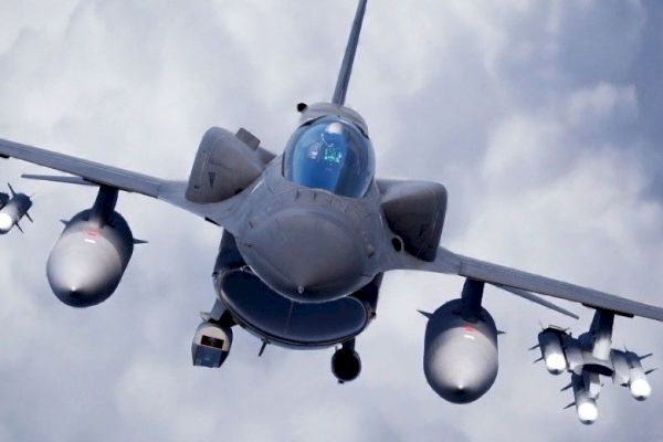 The image shows a front view of a military fighter jet flying with clouds in the background, equipped with various weapons.