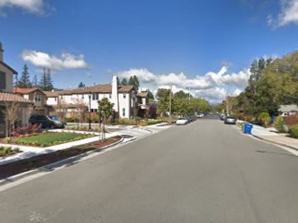 The image shows a suburban street with houses, parked cars, trees, and a blue trash bin on a clear day with a blue sky and some clouds.