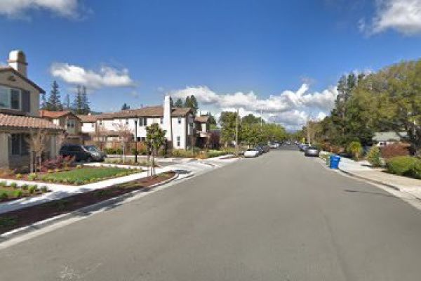 The image shows a suburban street with houses, parked cars, trees, and a blue trash bin on a clear day with a blue sky and some clouds.