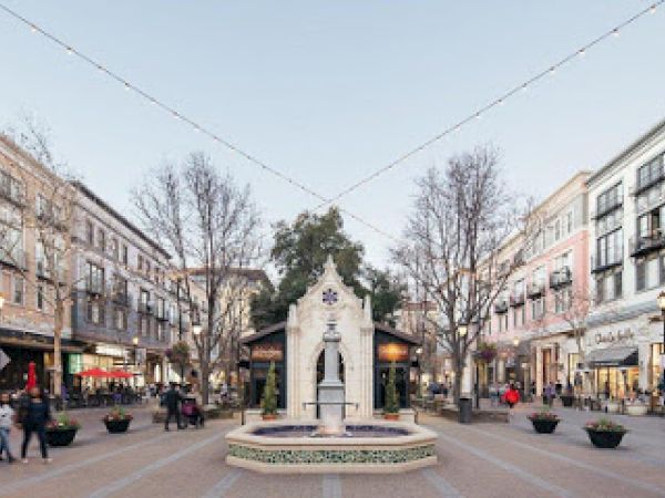 A bustling city street scene with people walking, surrounded by shops and restaurants, featuring a central fountain and overhead string lights.