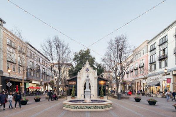 A bustling city street scene with people walking, surrounded by shops and restaurants, featuring a central fountain and overhead string lights.