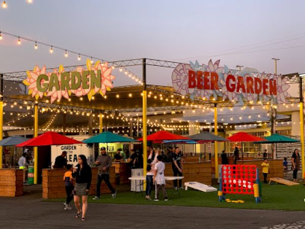 The image shows an outdoor beer garden with string lights, colorful umbrellas, people, and games like Connect Four under a sign that reads "BEER GARDEN."