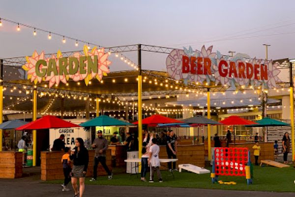 The image shows an outdoor beer garden with string lights, colorful umbrellas, people, and games like Connect Four under a sign that reads 