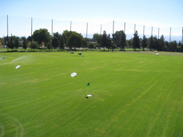 This image shows a well-maintained driving range with green grass, multiple targets, and surrounding trees under a clear blue sky.