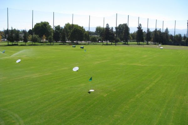 This image shows a well-maintained driving range with green grass, multiple targets, and surrounding trees under a clear blue sky.