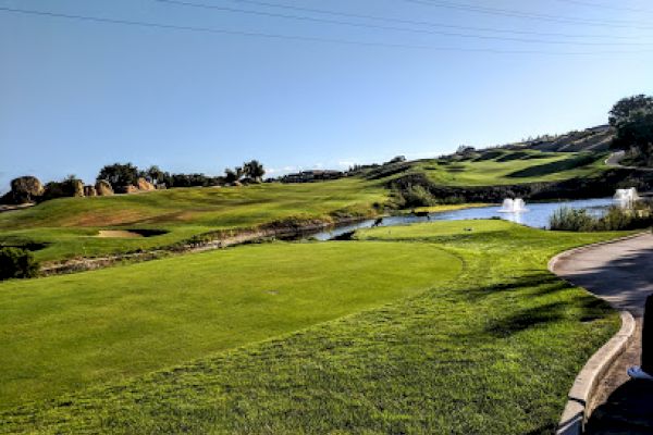 A well-maintained golf course with a lush green field, small water bodies, and fountains in a serene, sunny setting in an open landscape.