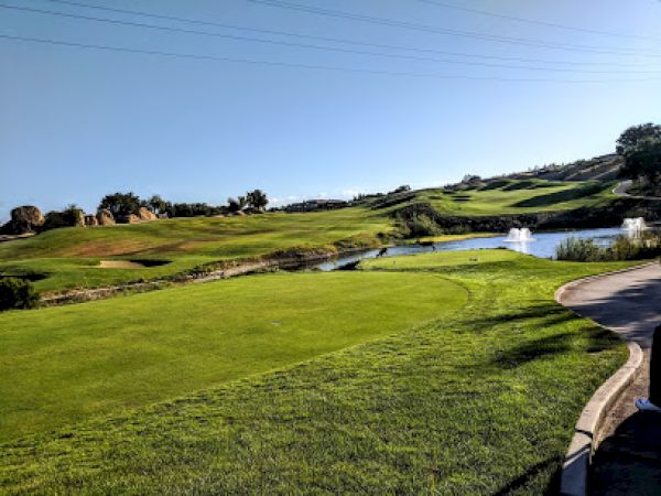A well-maintained golf course with a lush green field, small water bodies, and fountains in a serene, sunny setting in an open landscape.