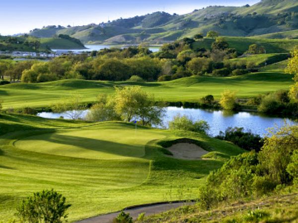 A scenic view of a golf course with rolling green hills, a small pond, and trees, under a clear sky with distant mountains.