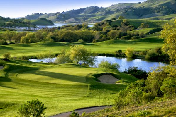 A scenic view of a golf course with rolling green hills, a small pond, and trees, under a clear sky with distant mountains.