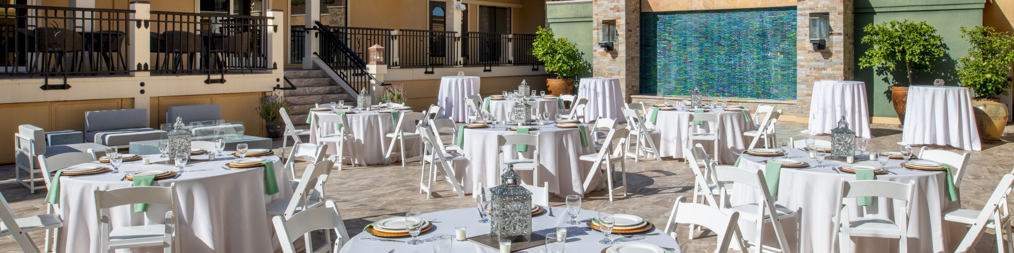 An outdoor patio is set for a formal event with round tables, white tablecloths, and white chairs against a backdrop of a multi-story building and blue sky.