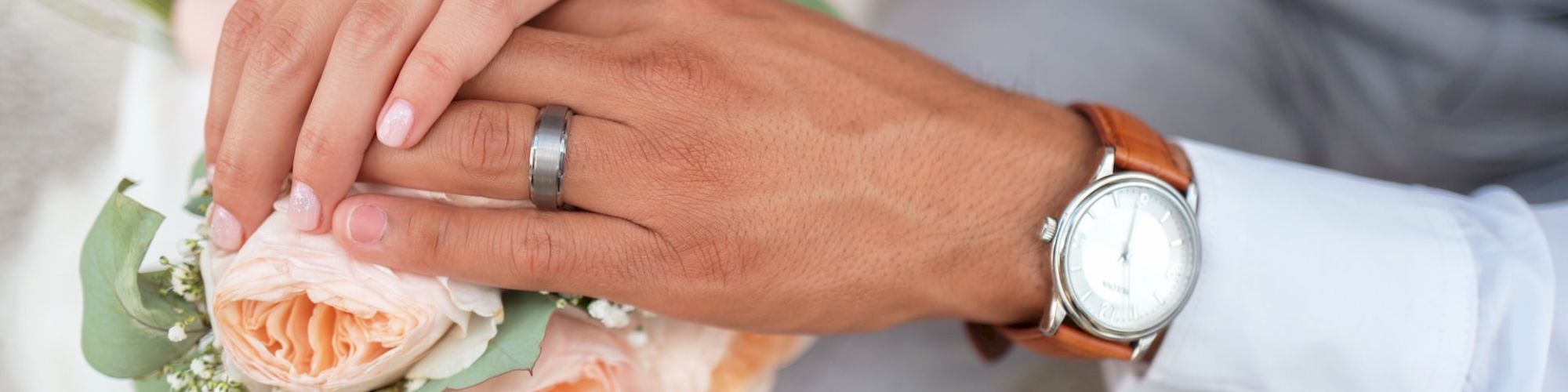 A bride and groom's hands are shown, with wedding rings visible, resting on a bouquet of peach roses and baby's breath.