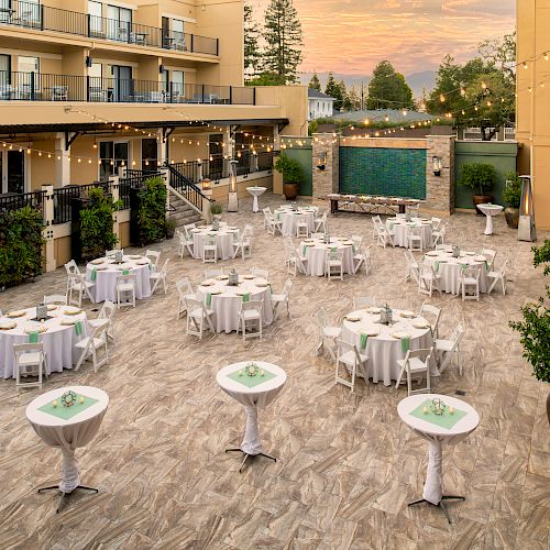 An outdoor courtyard with round and tall tables set up for an event, string lights overhead, and potted plants around, near a stone fountain wall.